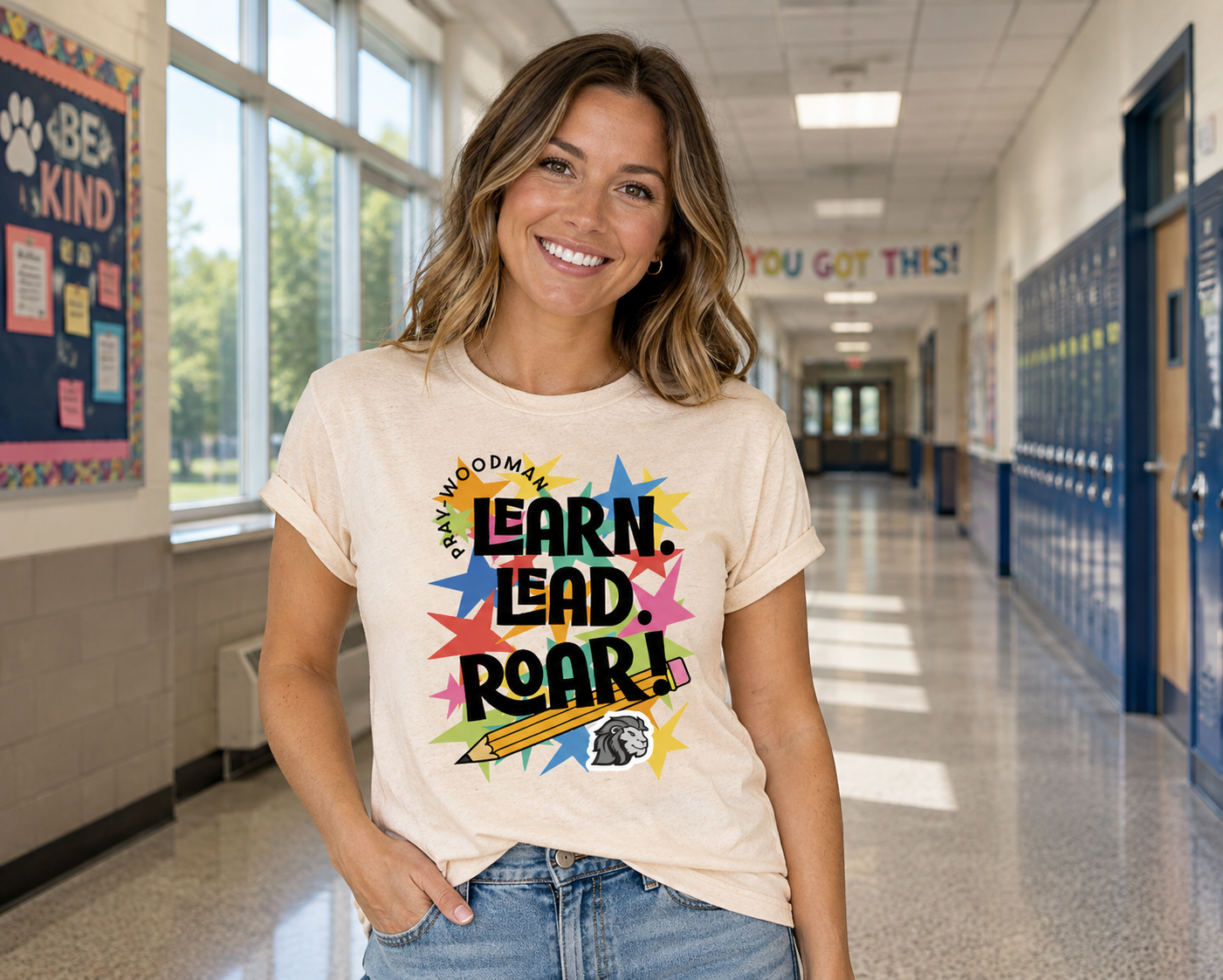 A woman smiling in a school hallway wearing a cream-colored graphic t-shirt with the text 'Pray-Woodman LEARN. LEAD.