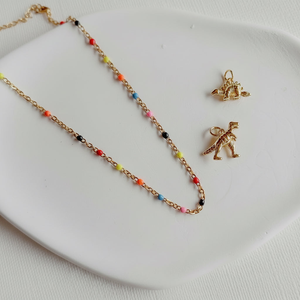 Gold necklace with colorful beads and two gold dinosaur pendants on a white plate.