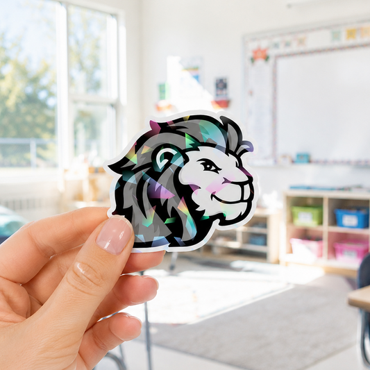 A hand holds a die-cut sticker featuring a stylized lion head with a holographic, iridescent foil finish, set against a blurred classroom background.